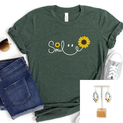 A dark green heather t-shirt with a white smiley face graphic and yellow sunflower details, styled in a flat lay with blue jeans, sunglasses, a white sneaker, and an inset showing matching sunflower earrings.