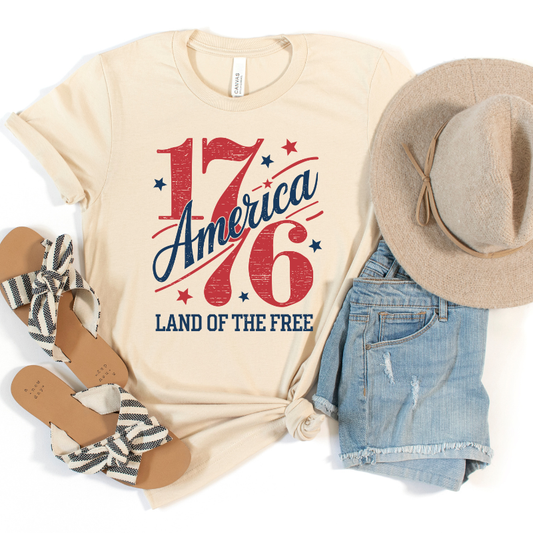 Soft cream t-shirt with 'America 1776 Land of the Free' design, paired with denim shorts, sandals, and a hat on a white background.