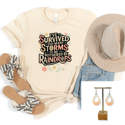 A styled flat-lay featuring a cream graphic T-shirt with the text 'I've survived too many storms to be bothered by raindrops' in a retro font, paired with a beige wide-brim hat, blue jeans, black-and-white striped sandals, and peach teardrop earrings on a stand.