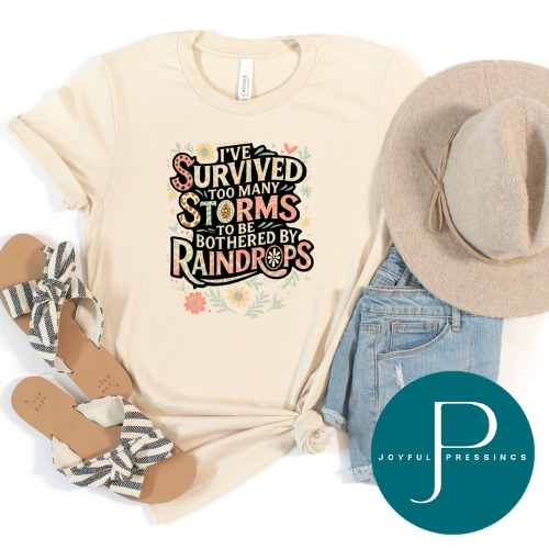 A styled flat-lay of a cream graphic T-shirt with the text 'I've survived too many storms to be bothered by raindrops' in a decorative multicolor font. The shirt is arranged with a beige wide-brim hat, light blue denim shorts, black-and-white striped sandals, and a teal circular 'Joyful Pressings' logo watermark on a white background.