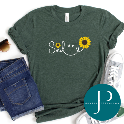 A dark heather green graphic t-shirt featuring a white cursive 'Smile' design with sunflower details, styled in a flat lay with blue denim shorts, sunglasses, white sneakers, and a teal circular 'JOYFUL PRESSINGS' brand watermark overlay.