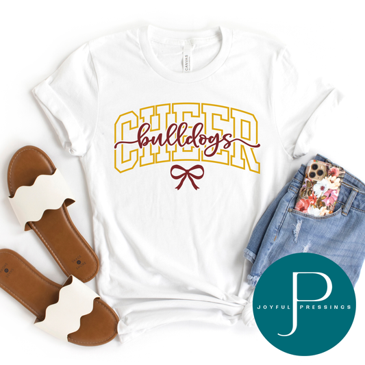 A white short-sleeve graphic T-shirt laid flat with denim shorts, brown slide sandals, and a floral phone case around it. The shirt has a large yellow outlined 'CHEER' text across the chest, a burgundy script 'bulldogs' in the center, and a burgundy bow graphic below.