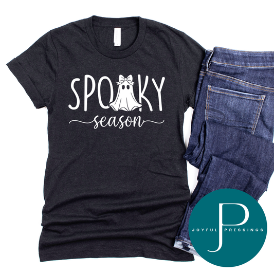 Black heather t-shirt with a white "Spooky Season" design, a cute ghost wearing a bow in place of the second O, laid flat beside folded dark jeans.
