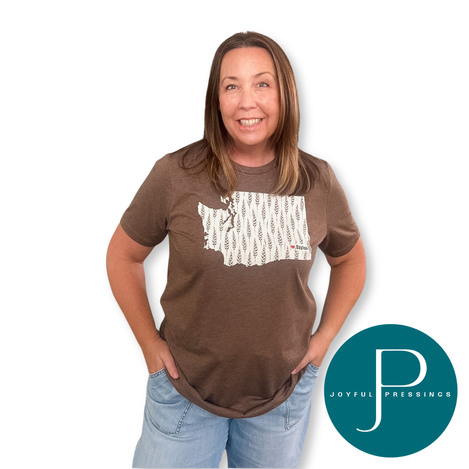 A woman modeling a brown short-sleeve t-shirt with a white and gray patterned graphic of the state of Washington on the front, paired with light blue jeans. A teal circular 'JOYFUL PRESSINGS' logo is in the bottom right corner against a plain white background.