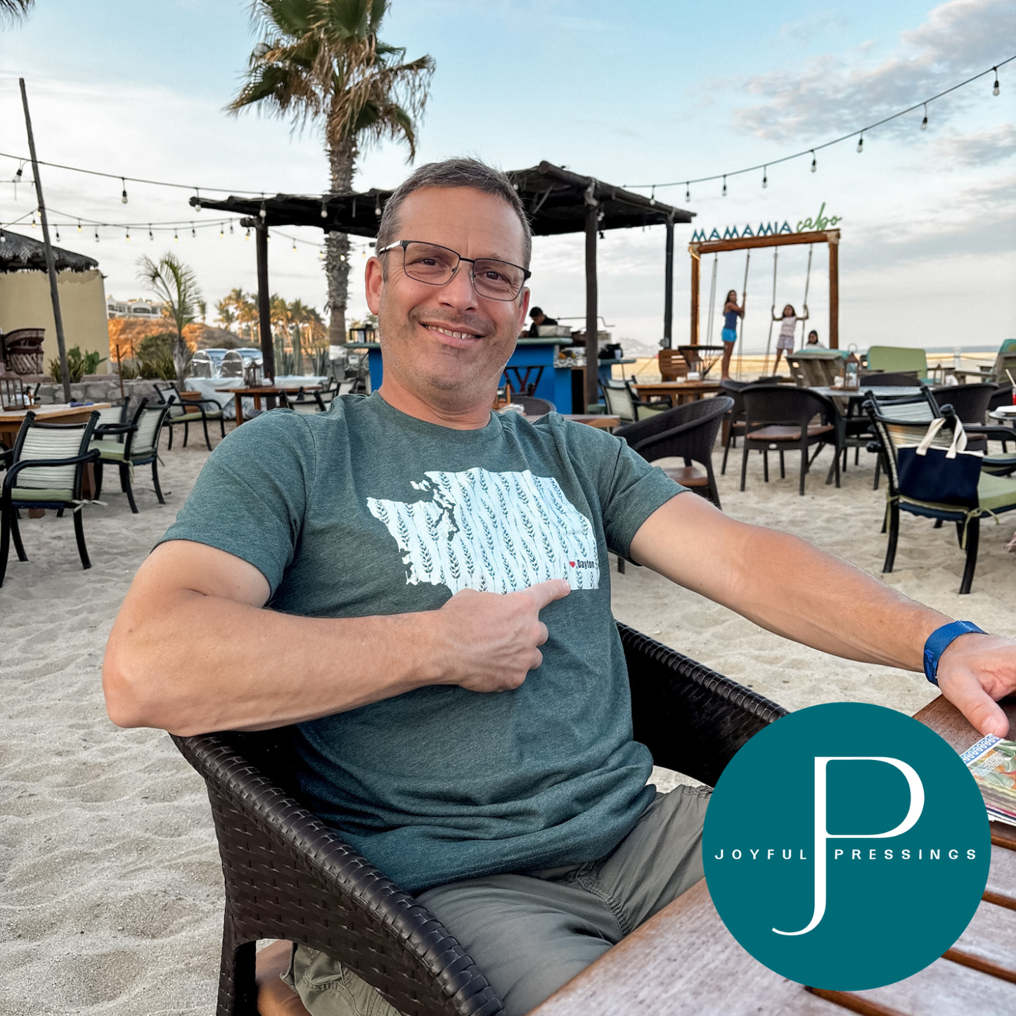A man wearing glasses and a green graphic t-shirt sits in a beachside patio chair, pointing toward the text on his shirt. The background shows a sandy beach, lounge chairs, and a palm tree under a cloudy sky.