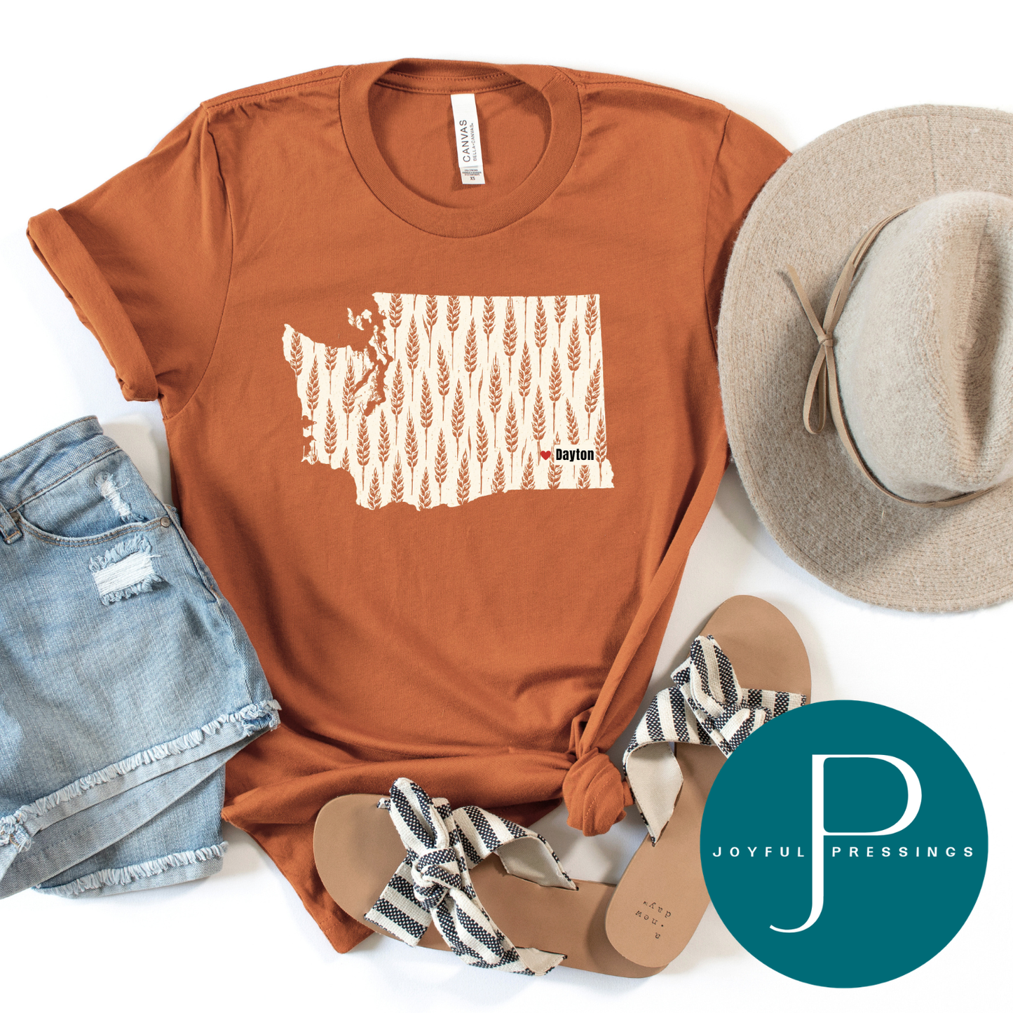 A rust-orange graphic t-shirt featuring a Washington state silhouette filled with a beige wheat pattern and the text 'Dayton'. The shirt is styled in a flat lay with a gray wide-brim hat, denim shorts, black and white patterned sandals, and a teal circular 'Joyful Pressings' logo overlay.