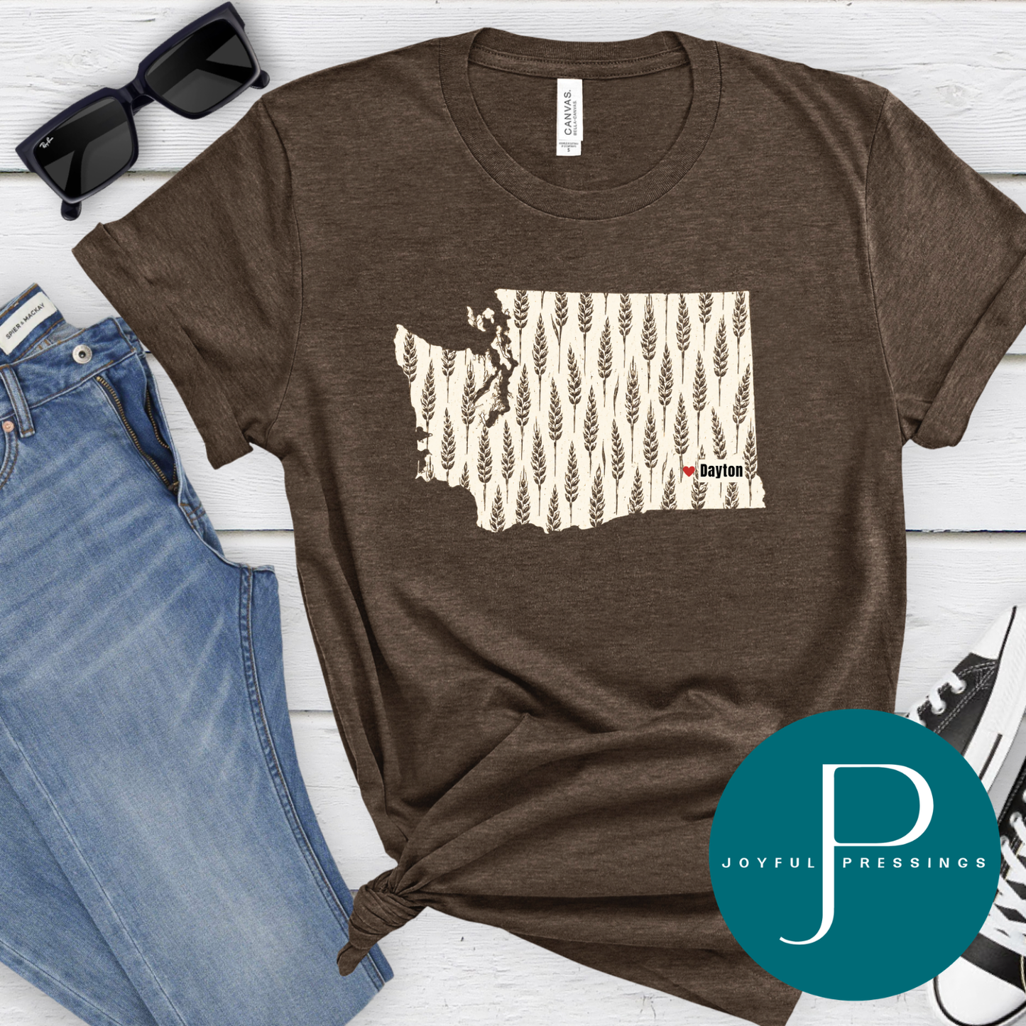 A brown heathered graphic T-shirt laid flat on a white wood background, styled with blue jeans, black sunglasses, and a black-and-white sneaker. The shirt has a cream map-of-Washington graphic filled with wheat stalks and small red text reading 'Dayton'.