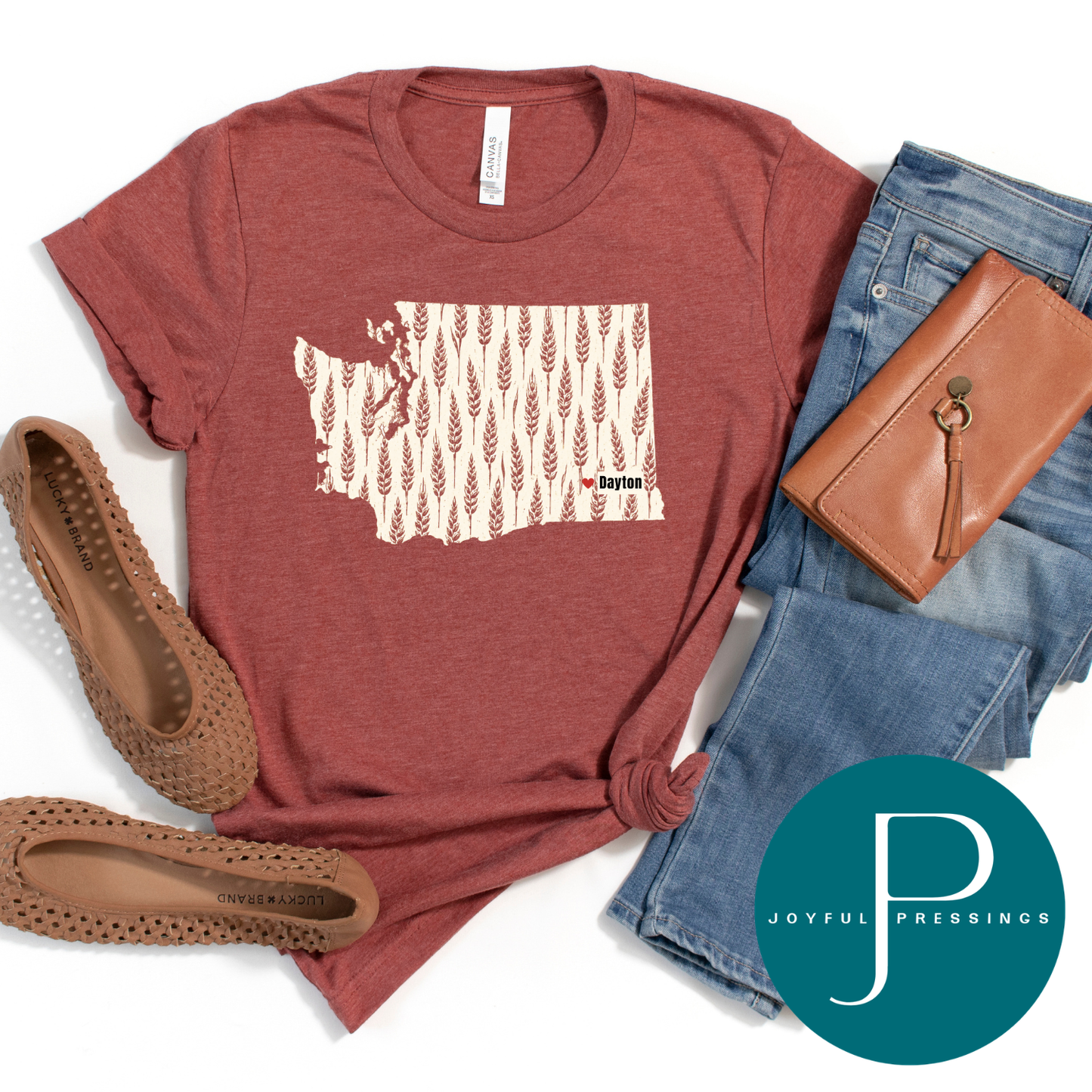 A styled flat-lay of a heathered rust-red graphic T-shirt with a cream and brown wheat pattern inside the outline of Washington state and the text 'Dayton'. The shirt is styled with blue jeans, a brown leather wallet, brown woven flats, and a teal circular 'Joyful Pressings' logo overlay on a white background.