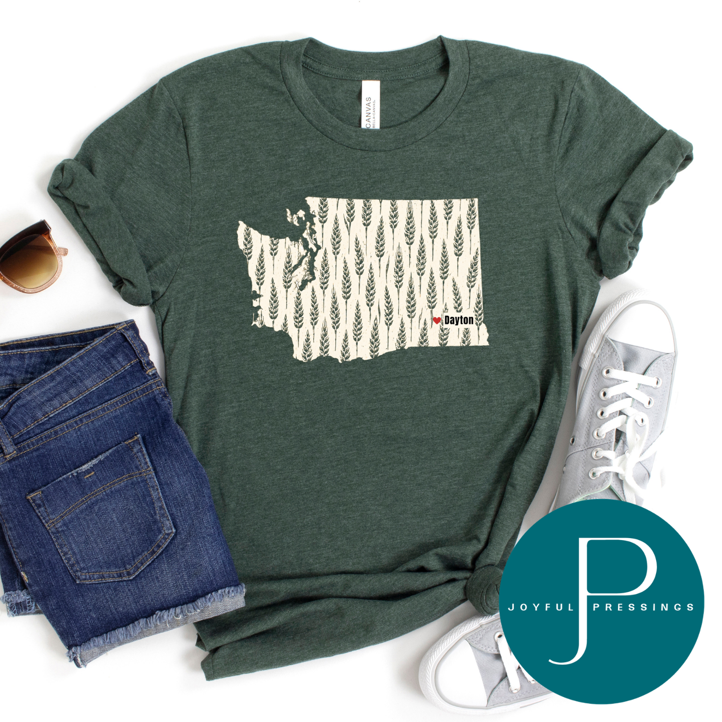 A dark heather green graphic t-shirt featuring a map of Washington filled with a wheat pattern and 'Dayton' text, styled in a flat lay with blue jeans, sunglasses, a white sneaker, and a teal circular 'Joyful Pressings' logo overlay.