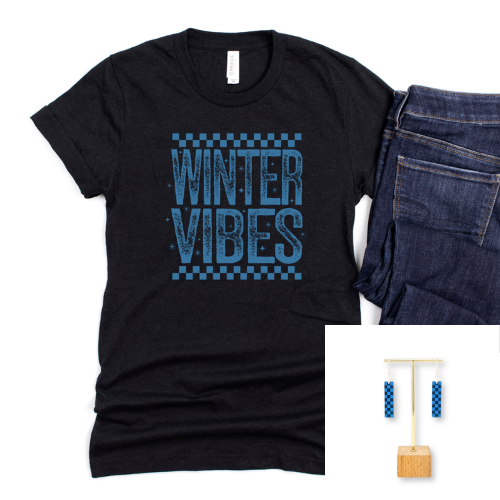 A black short-sleeve graphic T-shirt with blue distressed text reading 'WINTER VIBES' and a blue checker pattern, shown laid flat next to folded dark blue jeans on a white background. A small inset in the lower right shows blue patterned rectangular earrings on a wooden display stand.