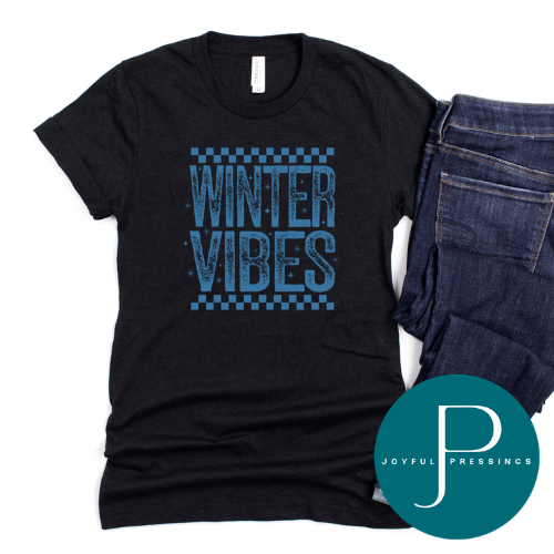 A black short-sleeve t-shirt with a blue distressed graphic reading 'WINTER VIBES' between checkerboard lines, styled flat-lay next to folded blue jeans on a white background. A teal circular 'JOYFUL PRESSINGS' watermark is in the bottom right corner.