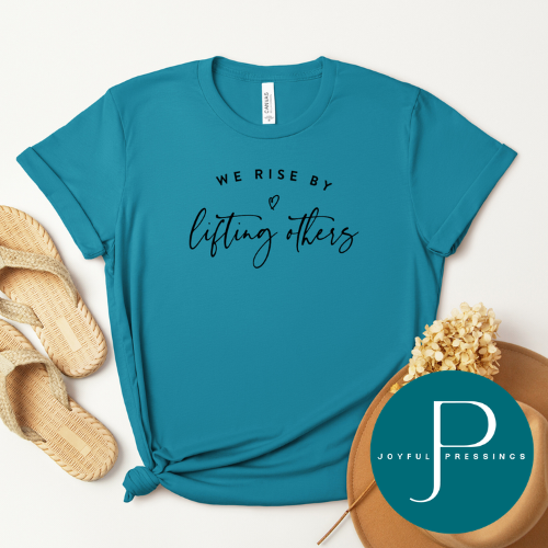 A teal short-sleeve graphic T-shirt laid flat on a light neutral background, styled with straw sandals, dried flowers, and a round branded Joyful Pressings badge. The shirt has black text reading 'WE RISE BY lifting others.