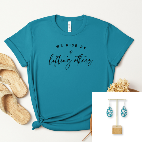 A teal short-sleeve graphic T-shirt laid flat on a light background, styled with woven sandals, dried flowers, and an inset showing matching teal teardrop earrings. The shirt has black text reading 'WE RISE BY lifting others.