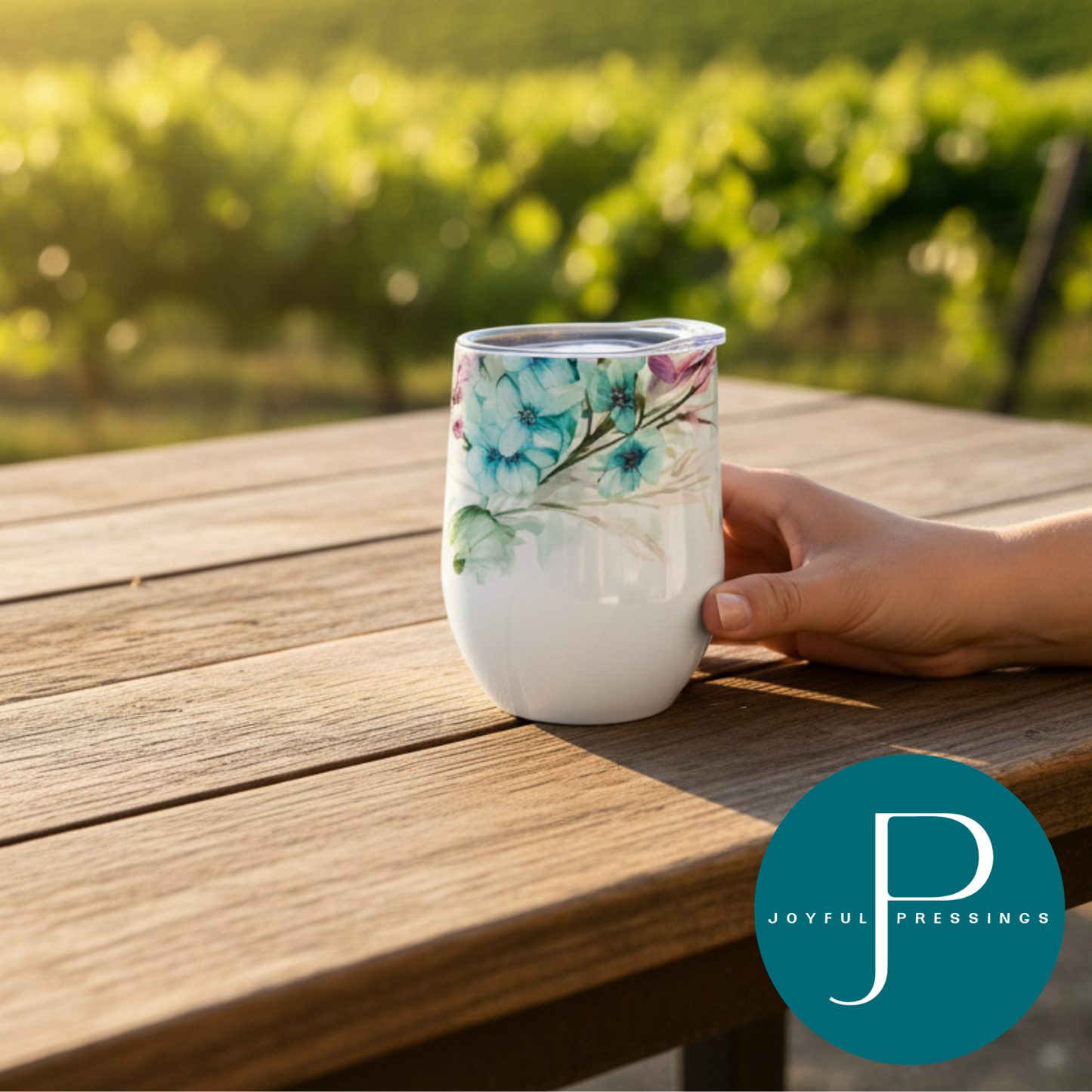 Hand holding a floral-patterned tumbler on a wooden table with vineyard background