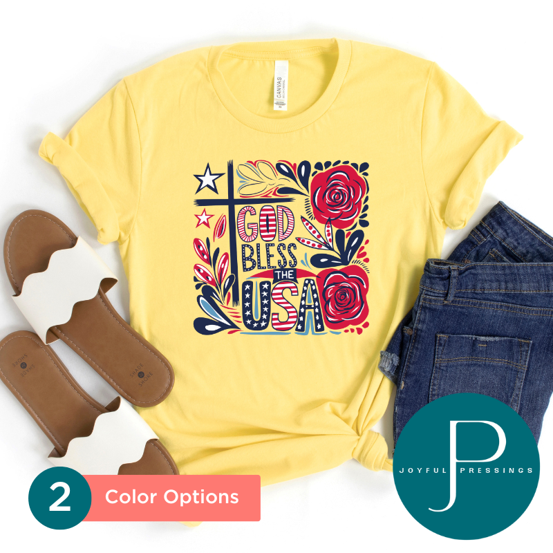 Flat lay of a yellow "God Bless the USA" t-shirt featuring a red, white, and navy floral cross design. Styled with denim jeans and white scalloped sandals.