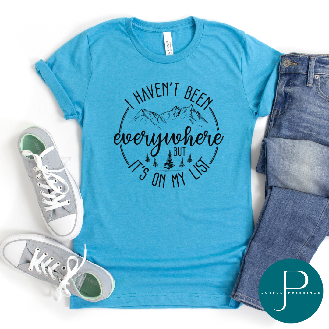 I Haven't Been Everywhere but it's on My List black print on a heather aqua shirt.  Displayed in a flatlay.