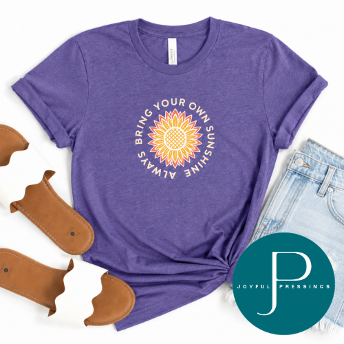 A heather purple graphic t-shirt featuring a sunflower design with the text 'BRING YOUR OWN SUNSHINE' in a circular layout. The t-shirt is styled in a flat-lay arrangement with light blue denim shorts, brown sandals, and a teal circular 'JOYFUL PRESSINGS' brand watermark overlay on a white background.