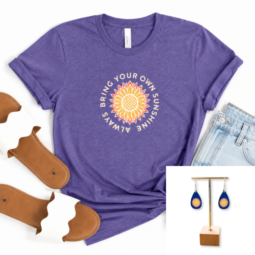 A heather purple graphic t-shirt featuring a sunflower design with the text 'BRING YOUR OWN SUNSHINE ALWAYS' in a circular layout. The t-shirt is styled in a flat-lay arrangement with light blue jeans, brown leather sandals, and an inset image showing blue teardrop earrings on a wooden stand.