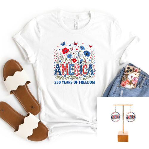 White graphic T-shirt laid flat with red, white, and blue floral artwork and the text 'AMERICA 250 YEARS OF FREEDOM.' Brown slide sandals, blue jeans with a floral pocket clip, and a small inset showing round earrings are arranged around it on a white background.
