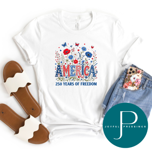 A white graphic T-shirt laid flat with decorative props including brown slide sandals, folded denim shorts with a floral pocket, and a teal circular Joyful Pressings logo overlay. The shirt graphic shows colorful flowers, butterflies, and the text 'AMERICA 250 YEARS OF FREEDOM.