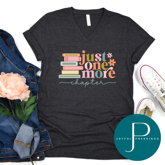 Dark grey heather v-neck t-shirt with colorful stacked books and the phrase ‘Just One More Chapter’ styled with denim jacket, sneakers, and a flower.