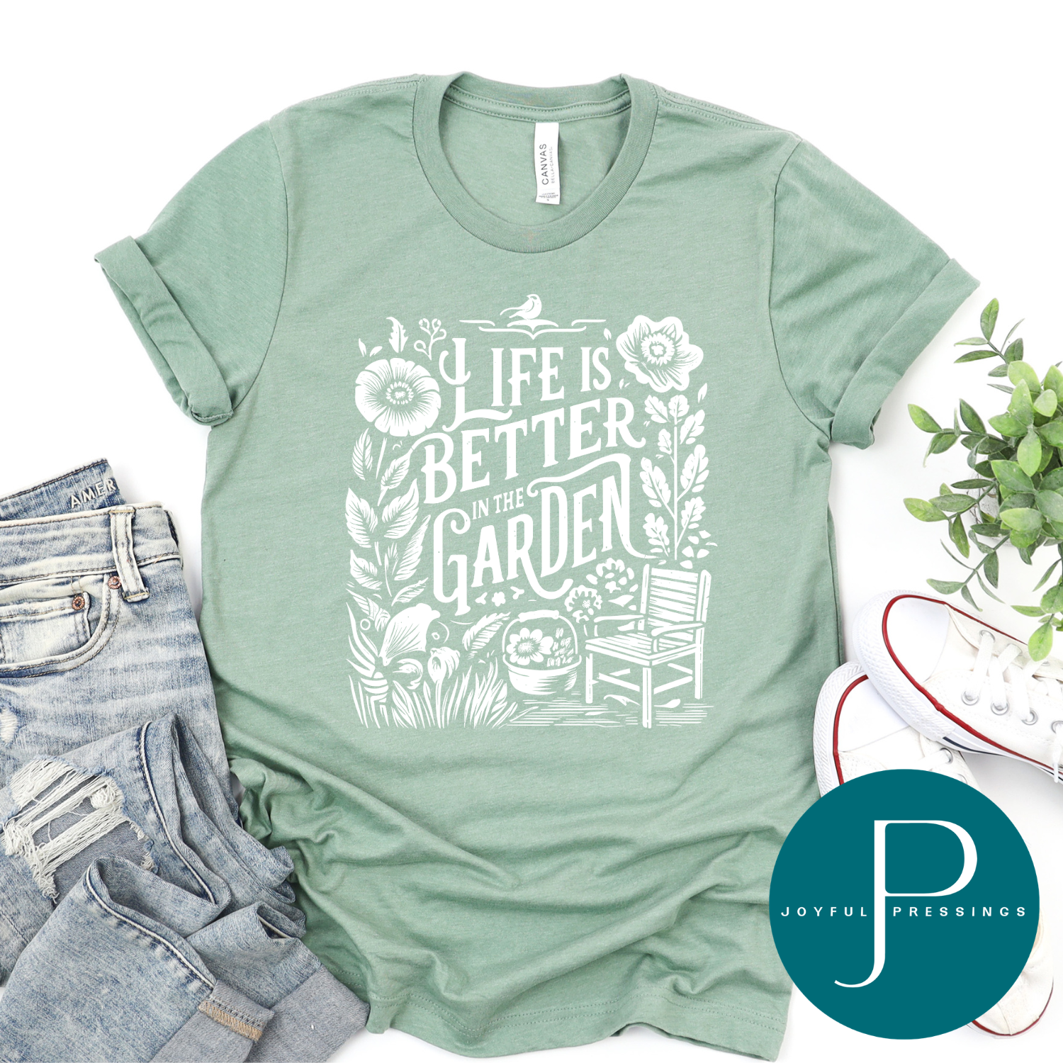 Heather sage green “Life Is Better in the Garden” Bella + Canvas t-shirt with a white floral and garden chair graphic, styled flat lay with jeans, white sneakers, greenery, and Joyful Pressings logo.