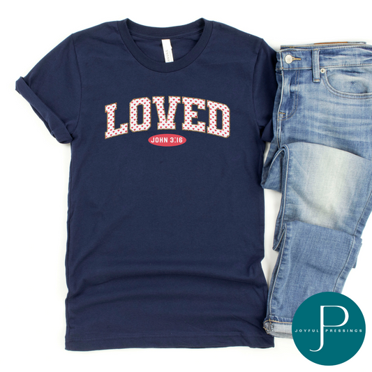 flatlay of navy blue tee with the word loved in white and adorned with pink hearts.  underneath is the reference to the verse John 3:16