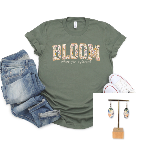 An olive green graphic t-shirt with 'BLOOM where you're planted' in floral letters, styled in a flat lay with blue jeans, white sneakers, and a pair of dangling earrings on a stand.