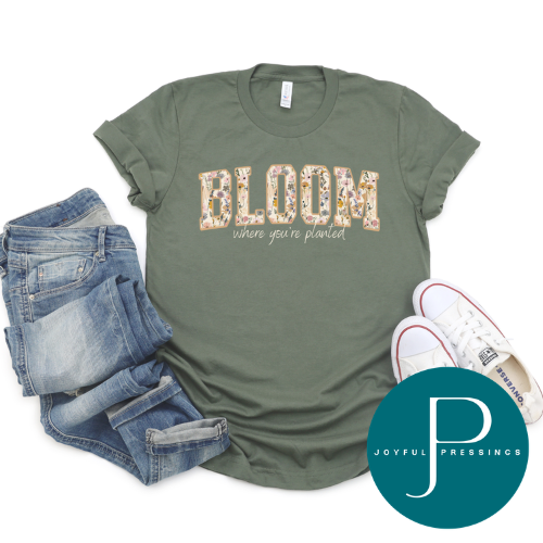 An olive green graphic t-shirt featuring the word 'BLOOM' in a floral patterned font and the phrase 'where you're planned' underneath. The shirt is styled in a flat lay with blue jeans, white sneakers, and a teal circular 'JOYFUL PRESSINGS' brand watermark on a white background.