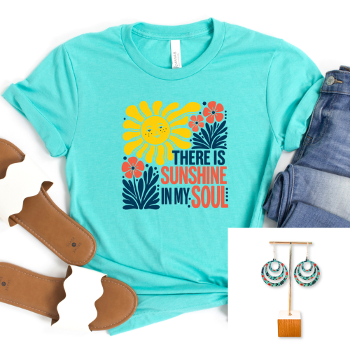 A turquoise short-sleeve graphic T-shirt laid flat on a white background, styled with folded blue jeans, brown leather accessories, and an inset showing matching turquoise earrings. The shirt has a colorful sunflower and floral graphic with the text 'THERE IS SUNSHINE IN MY SOUL.