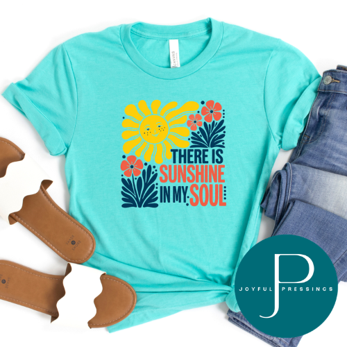 A turquoise short-sleeve graphic T-shirt laid flat with folded blue jeans, brown sandals, and a teal circular Joyful Pressings logo on a white background. The shirt has a colorful sun, flowers, and the phrase 'THERE IS SUNSHINE IN MY SOUL.
