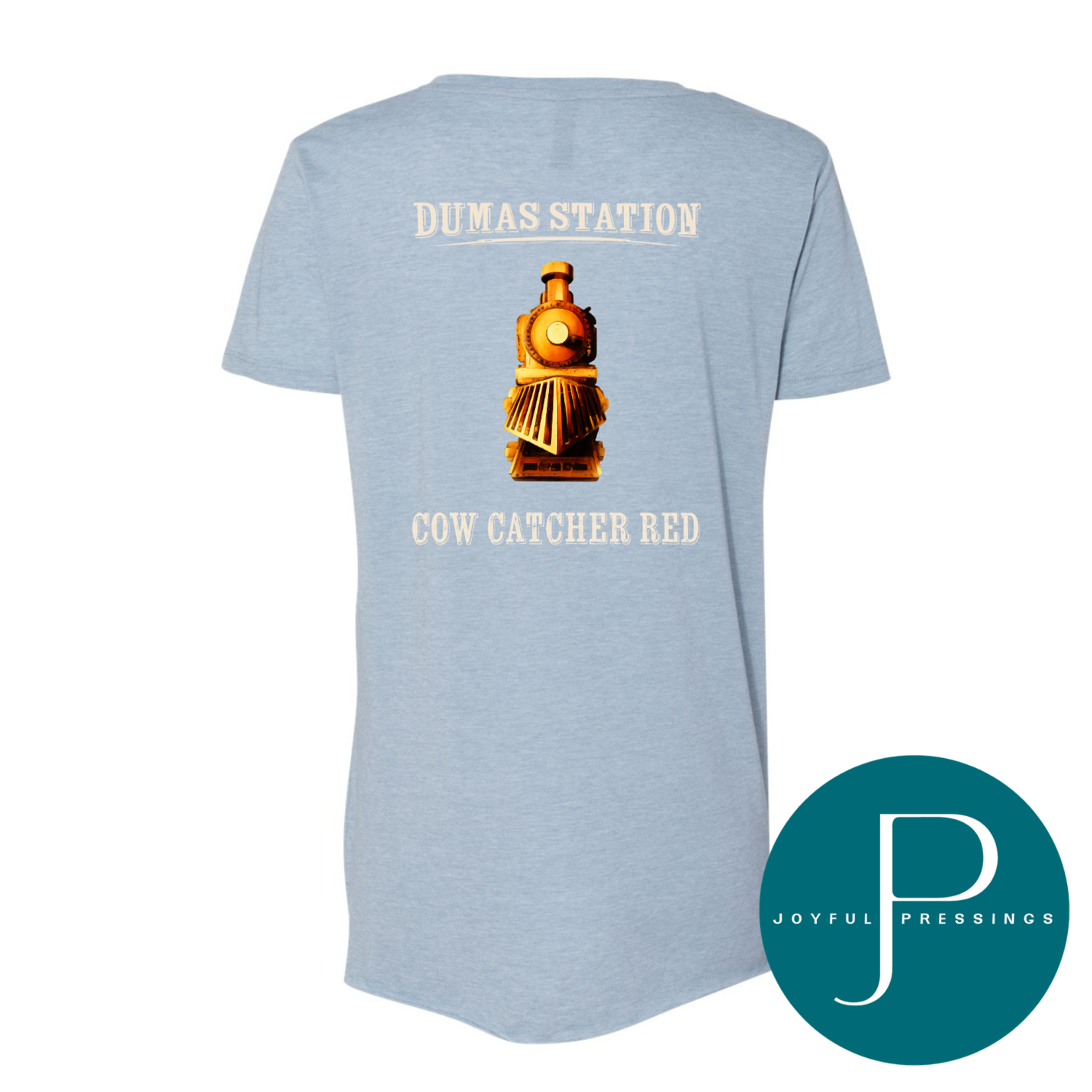 A light heather blue short-sleeve t-shirt shown from the back, featuring a vintage-style brown and gold train graphic with cream-colored text reading 'DUMAS STATION' above and 'COW CATCHER RED' below. A teal circular 'JOYFUL PREBRINGS' logo is in the bottom right corner.