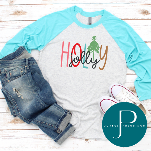 A light heather gray raglan baseball T-shirt with bright aqua sleeves, styled in a flat lay with blue jeans, white sneakers, and a teal circular Joyful Pressings logo on a white wood plank background. The shirt features a colorful 'HOLJOLY' graphic with a small green Christmas tree.
