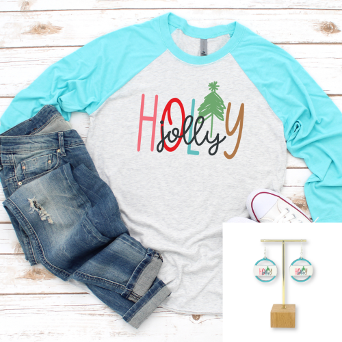 A light heather gray and turquoise raglan baseball t-shirt with 'HOLLY jolly' written across the chest in colorful lettering and a Christmas tree graphic, styled in a flat lay with blue jeans and a white sneaker on a white wood background. A small inset in the bottom right shows matching earrings on a display stand.