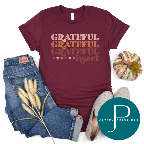 A flat-lay outfit arrangement featuring a maroon short-sleeve graphic T-shirt with the words 'GRATEFUL GRATEFUL GRATEFUL heart' in warm tan and brown lettering, styled with blue jeans, a small striped pumpkin, beige flats, and dried grass stems on a white background. A teal circular 'Joyful Pressings' logo appears in the lower right corner.