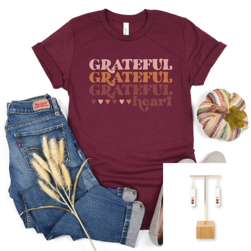 A flat-lay outfit arrangement featuring a maroon short-sleeve graphic T-shirt with the words 'GRATEFUL GRATEFUL GRATEFUL heart' in warm tan and brown tones, styled with blue jeans, a striped decorative pumpkin, dried grass stems, and a small inset showing dangling earrings.