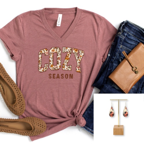A dusty rose V-neck t-shirt with 'COZY SEASON' printed in floral block letters, styled in a flat lay with blue jeans, a brown leather wallet, brown woven flats, and an inset showing matching floral teardrop earrings.