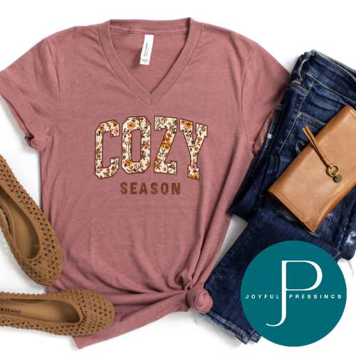 A dusty rose V-neck t-shirt with 'COZY SEASON' printed in floral block letters, styled in a flat lay with blue jeans, a brown leather wallet, brown woven flats, and a Joyful Pressings logo overlay.