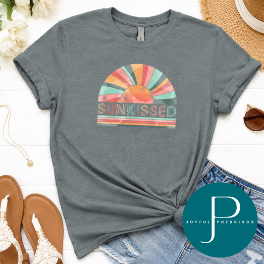 Heather Blue Storm t-shirt with colorful graphic and 'Sunkissed' text, placed on a white surface with denim jeans and sandals.