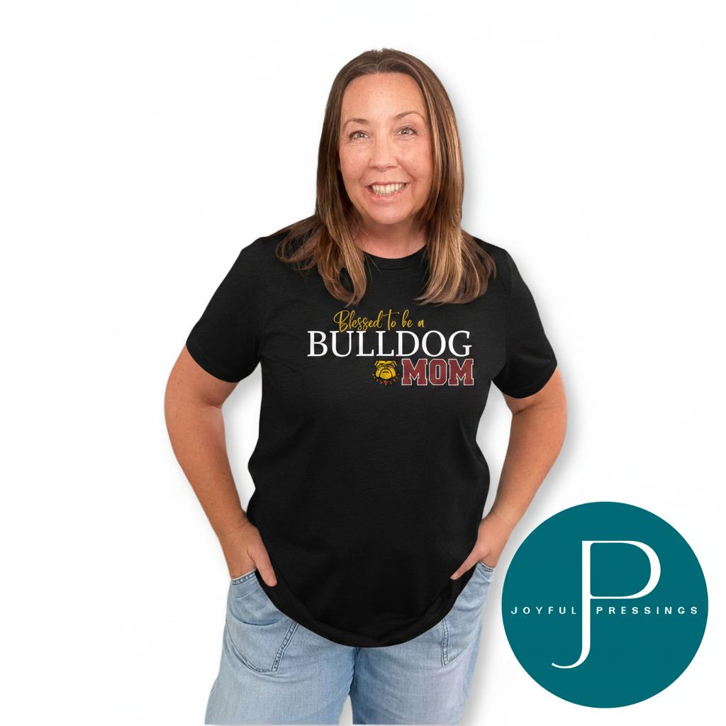A woman wearing a black short-sleeve graphic T-shirt with the text 'Blessed to be a BULLDOG MOM' in white, yellow, and red, paired with light blue jeans, against a plain white background. A teal circular 'JOYFUL PRESSINGS' logo is in the bottom right corner.