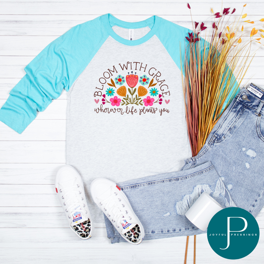 Next Level Tahiti Blue and Heather White raglan t-shirt with colorful floral graphic and the phrase “Bloom with Grace Wherever Life Plants You,” encouraging joy and growth in any season.