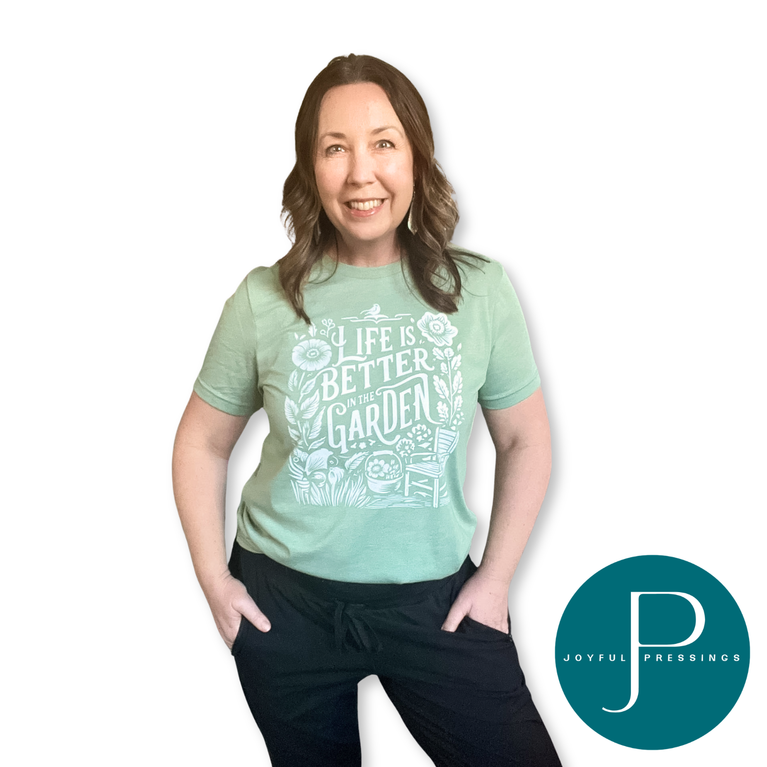 Smiling woman wearing a heather sage green “Life Is Better in the Garden” graphic t-shirt with a white floral and garden chair design, hands in pockets, on a white background with Joyful Pressings logo.