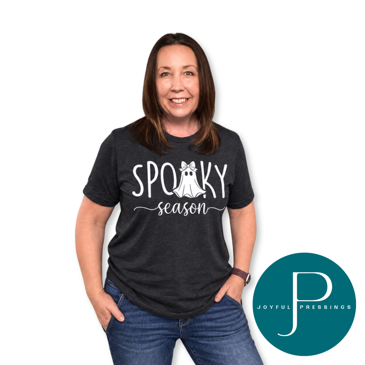 Smiling woman with shoulder-length brown hair wearing a black heather "Spooky Season" t-shirt with a bow-wearing ghost, styled with blue jeans.