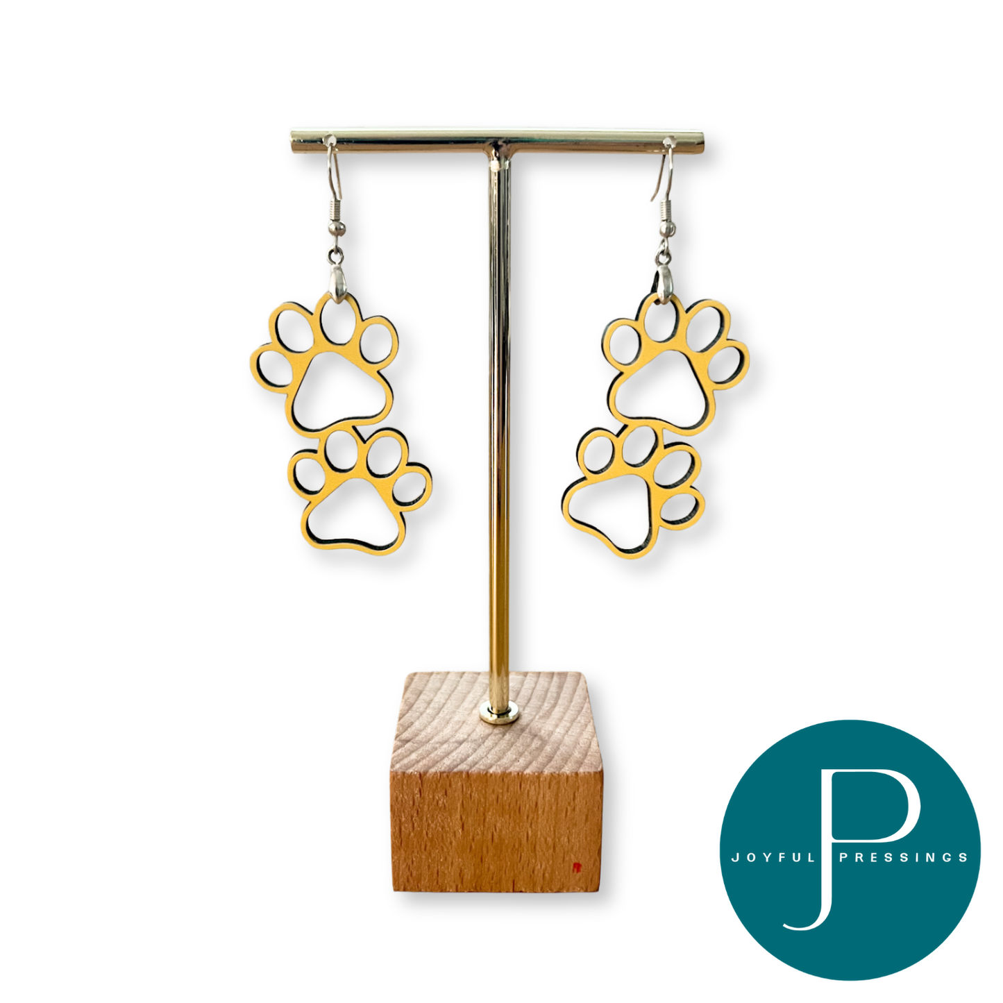Gold paw print dangle earrings displayed on a gold T-bar jewelry stand with a wooden base, set against a plain white background with a teal Joyful Pressince logo in the bottom right corner.