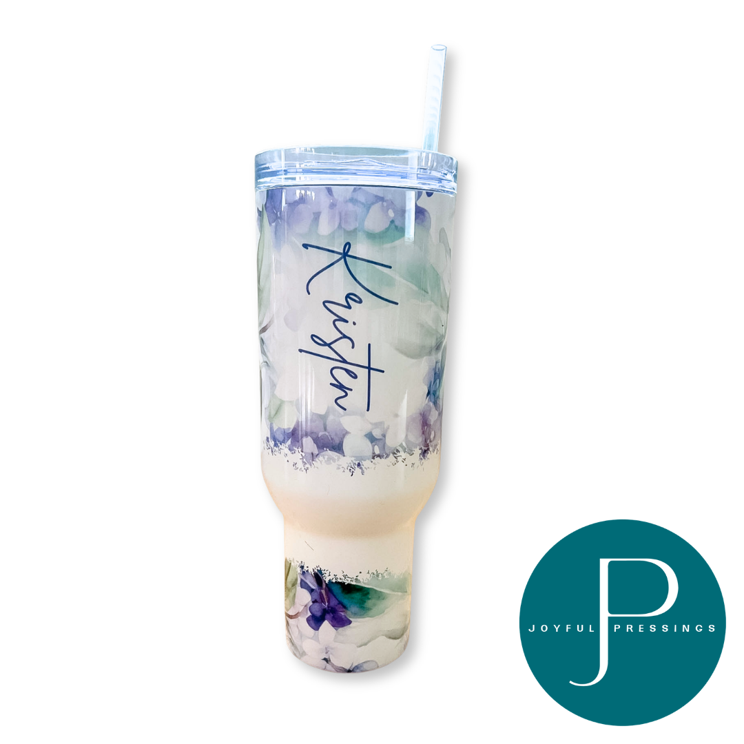A white insulated tumbler with a lid and straw, featuring a watercolor floral design in purple, blue, and green with a cream-colored lower section. The word 'Kisten' is printed in purple script on the front.