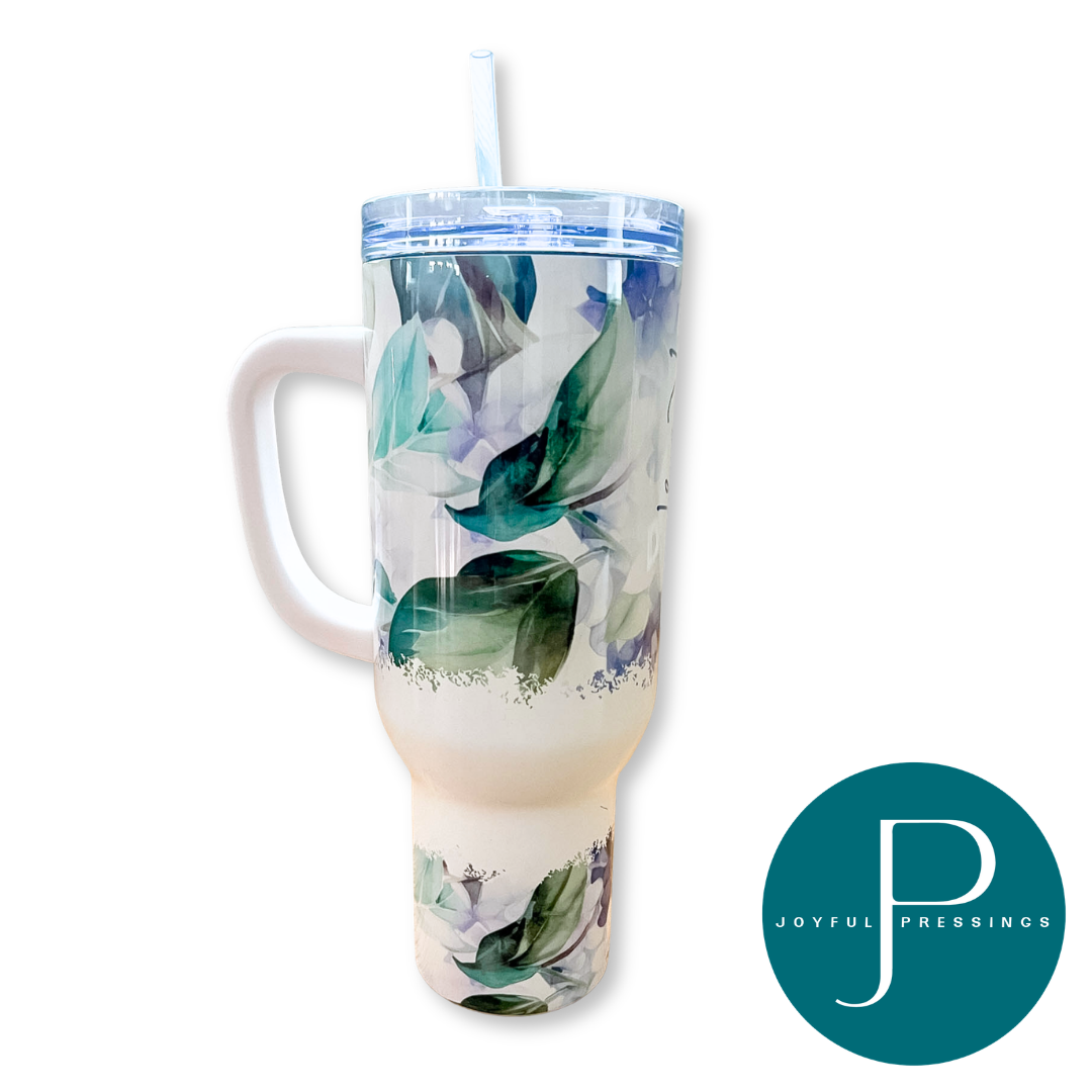 A tall handled tumbler with a clear lid and straw, featuring a watercolor floral print in shades of green, blue, and white on a clear base. A teal circular 'Joyful Pressings' logo is displayed in the bottom right corner against a solid white background.
