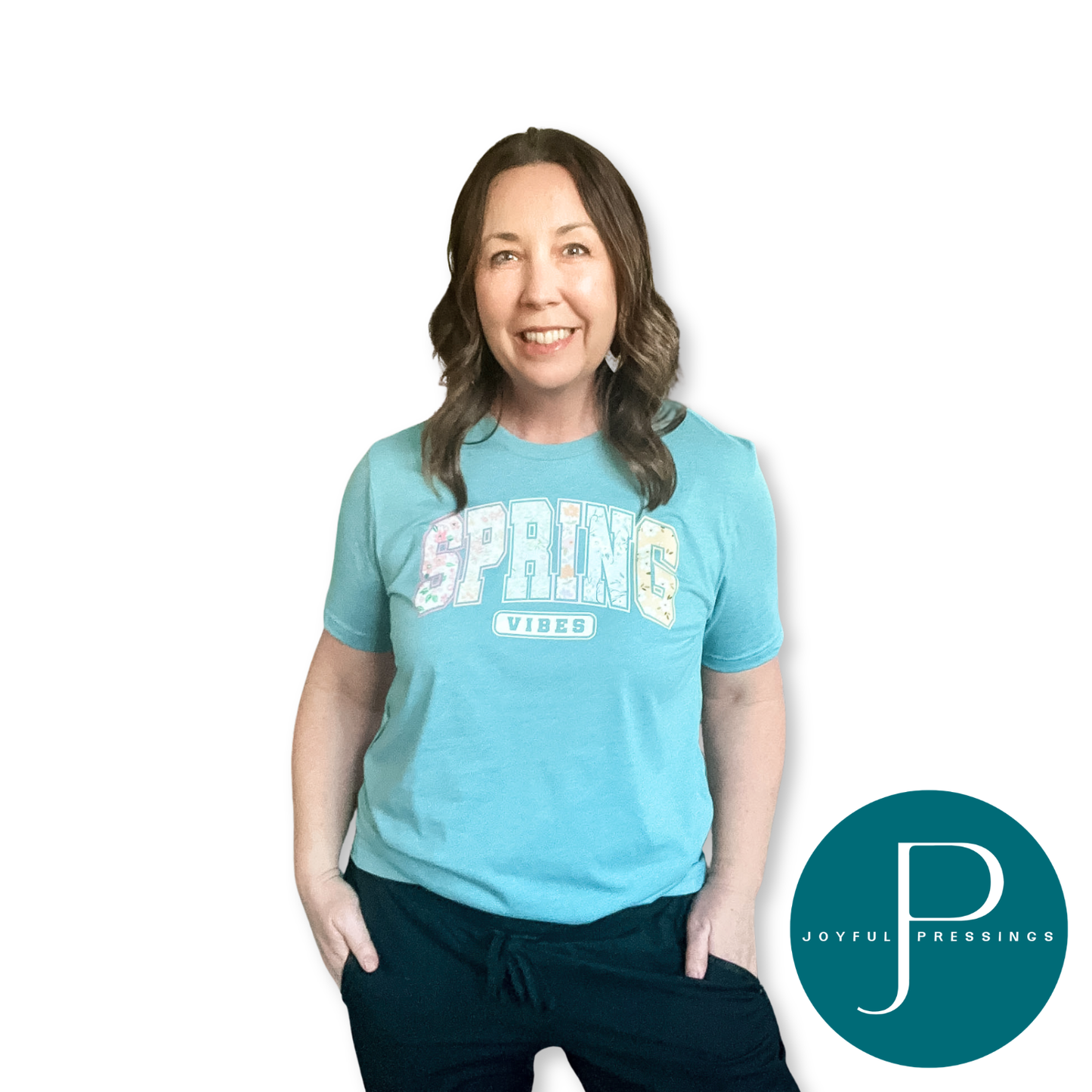 Smiling woman wearing a heather blue lagoon “Spring Vibes” graphic t-shirt with a colorful floral “SPRING” design and “VIBES” underneath, hands in pockets, on a white background with Joyful Pressings logo.