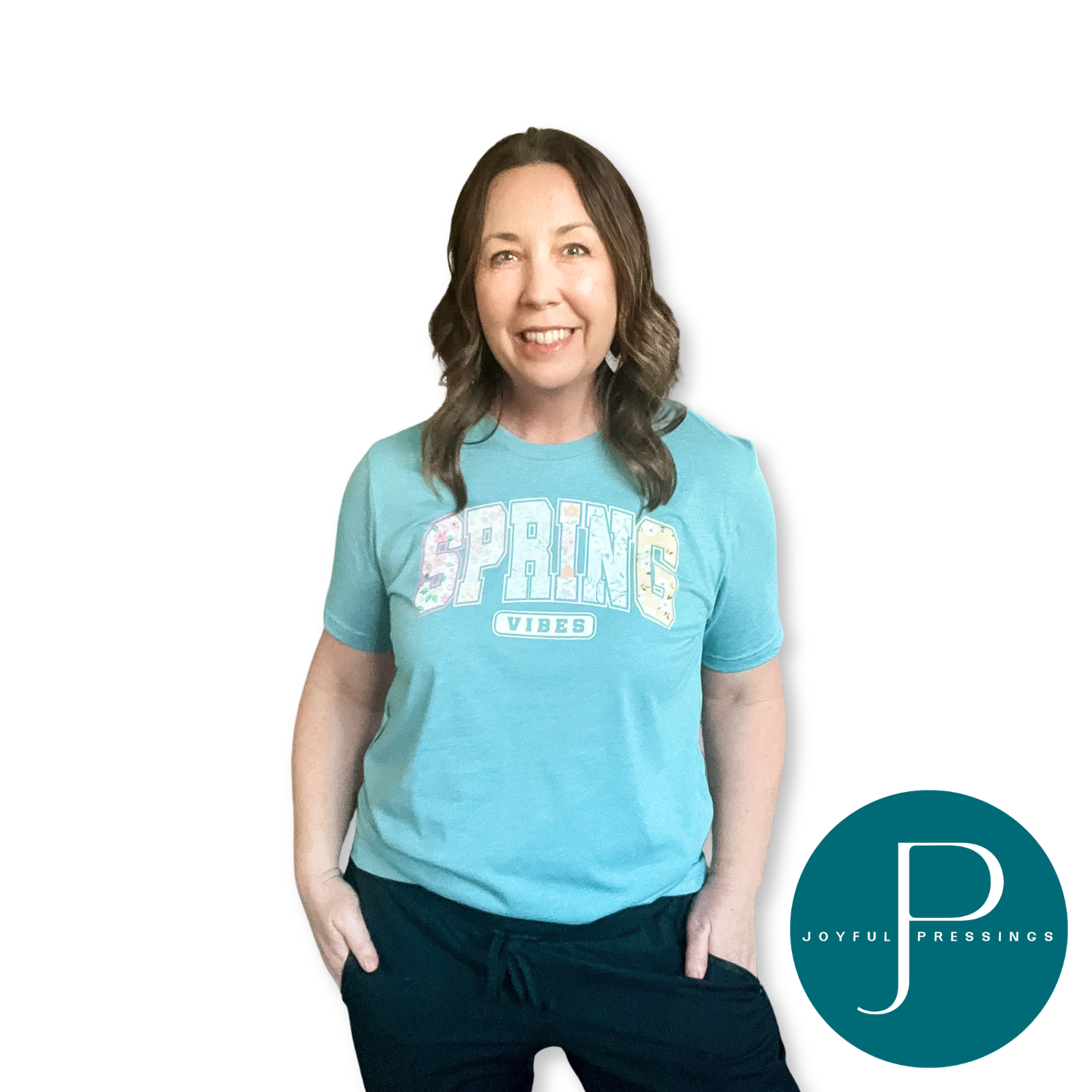 Smiling woman wearing a heather blue lagoon “Spring Vibes” graphic t-shirt with a colorful floral “SPRING” design and “VIBES” underneath, hands in pockets, on a white background with Joyful Pressings logo.