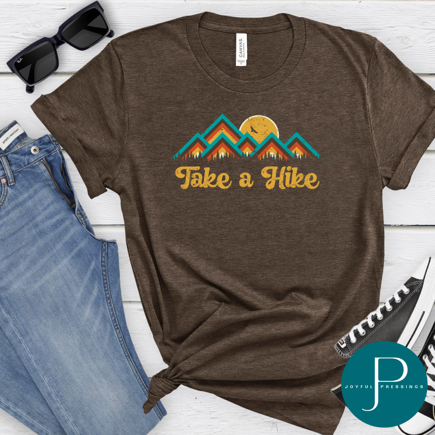 Flat lay of a heather brown t-shirt with a colorful retro mountain design and the words “Take a Hike,” styled with denim shorts, tan sandals, and a straw hat. 