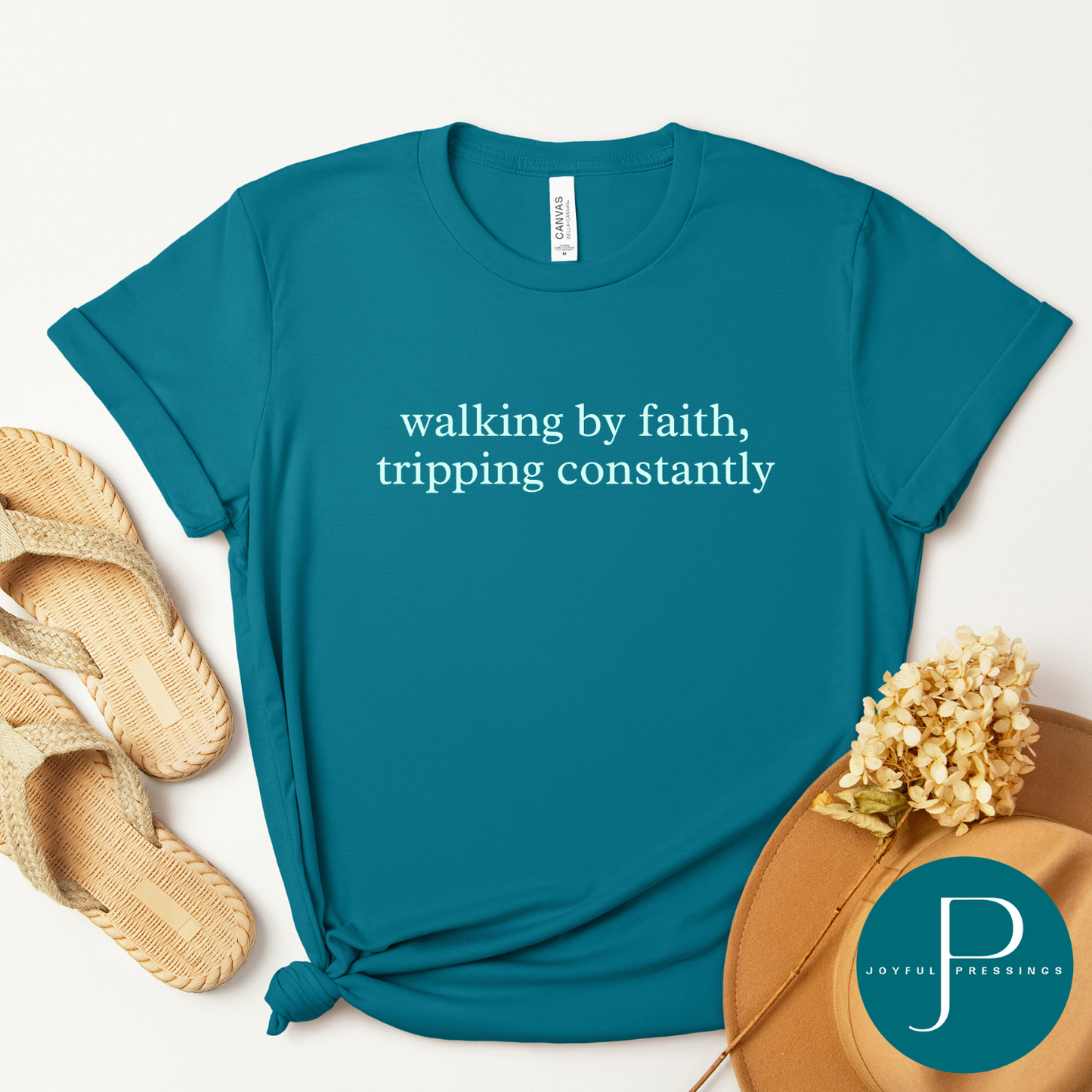 Flat lay of a marine blue t-shirt with the words “walking by faith, tripping constantly” in simple white text. Styled with neutral flip-flop sandals, a tan felt hat, and dried flowers.